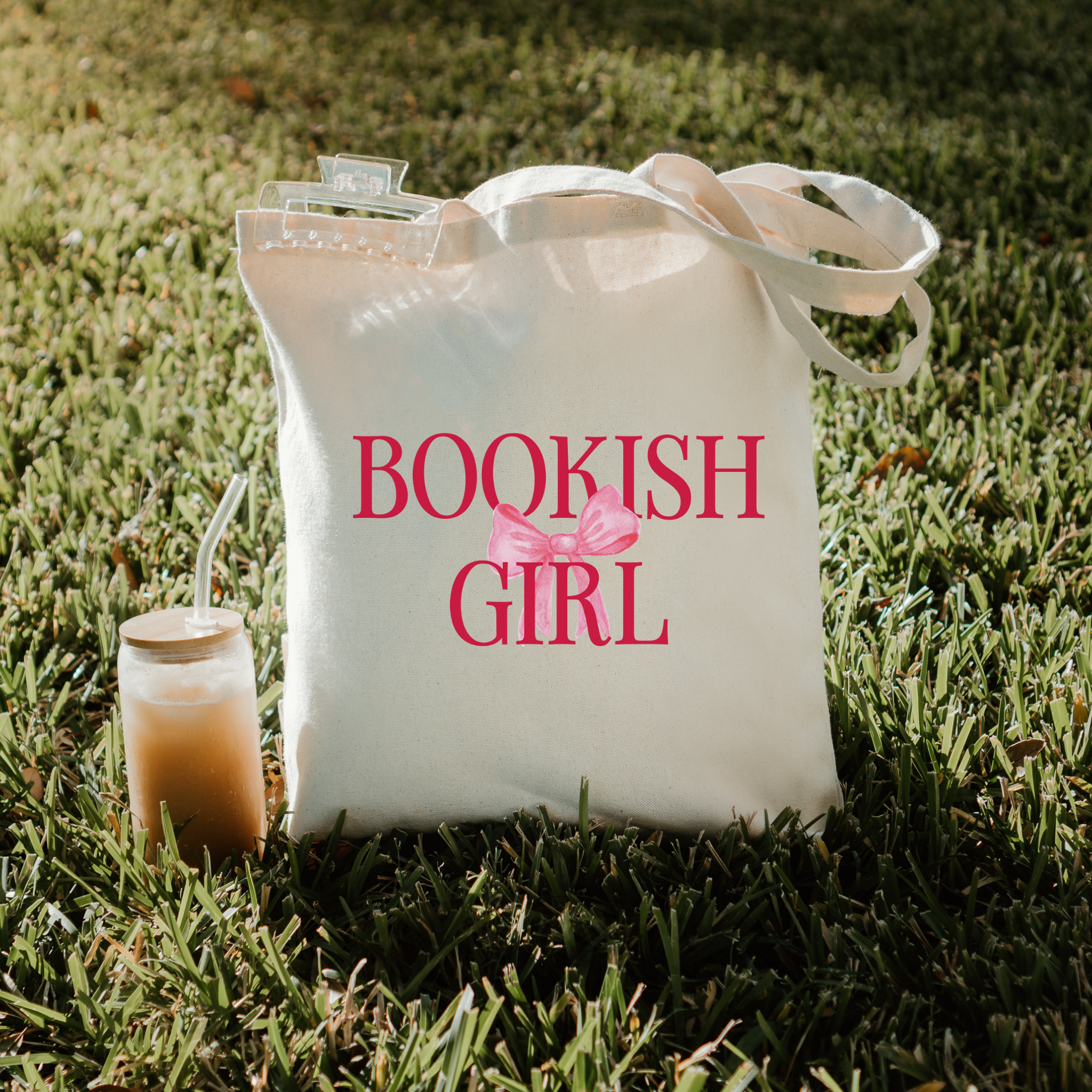 White tote bag with 'BOOKISH GIRL' text and a pink bow on grass