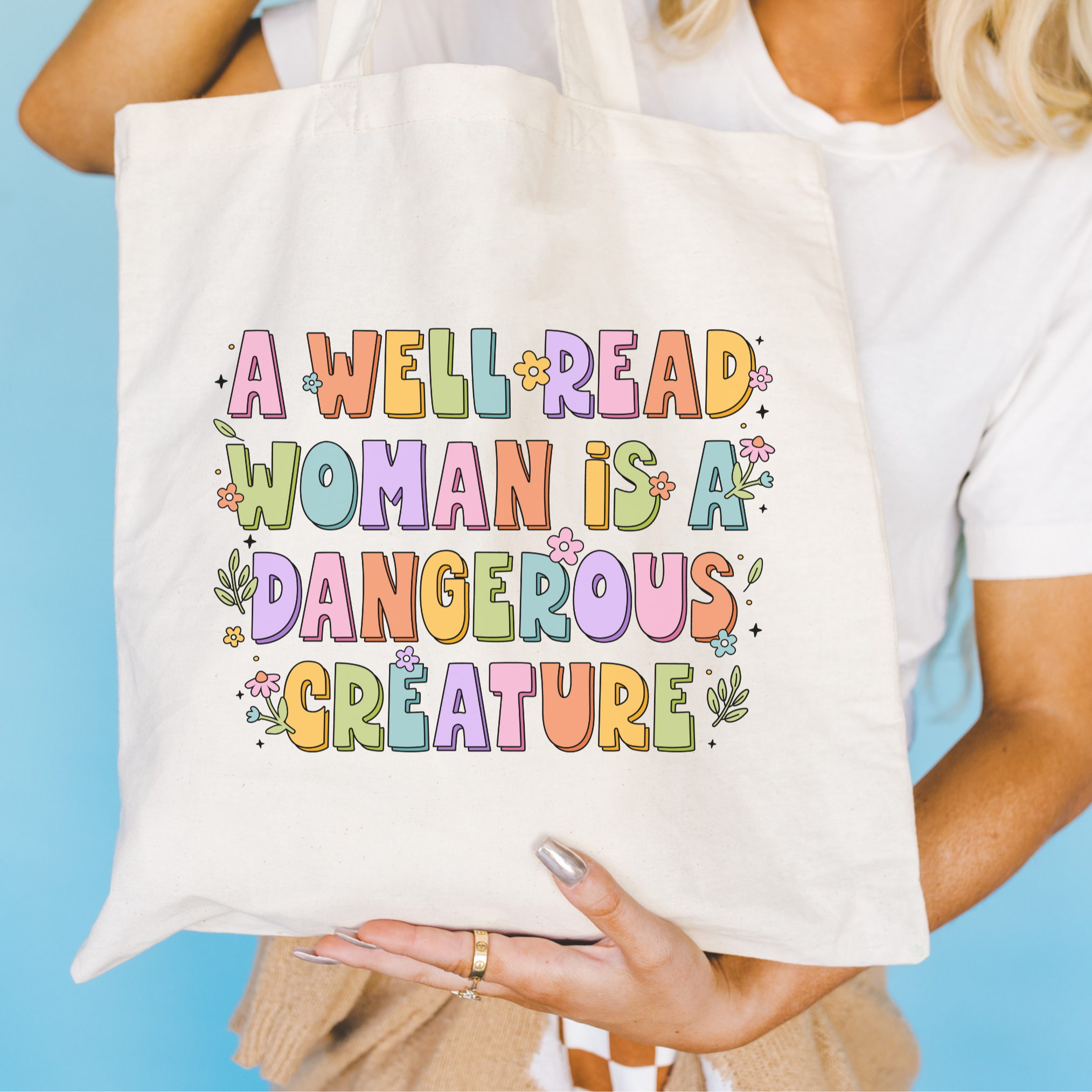 Tote bag with colorful text 'A Well Read Woman is a Dangerous Creature' held by a person against a blue background.
