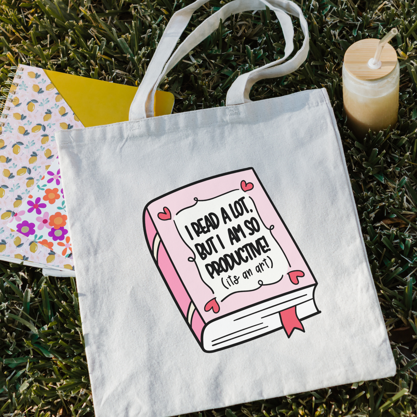 Bookish tote bag with a book graphic and text on grass with a drink and notebook.