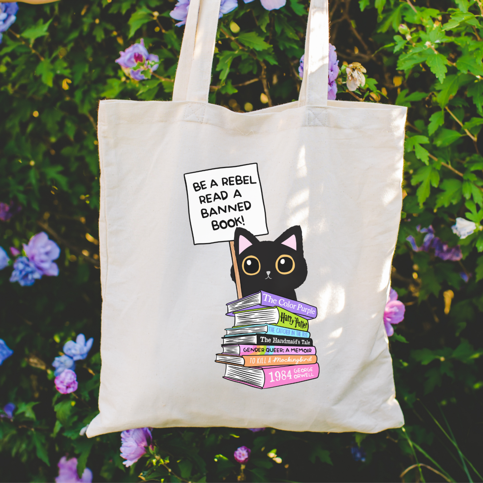 Bookish Tote bag with a cat and books design held in front of a floral background with text Be a Rebel and Read a Banned Book