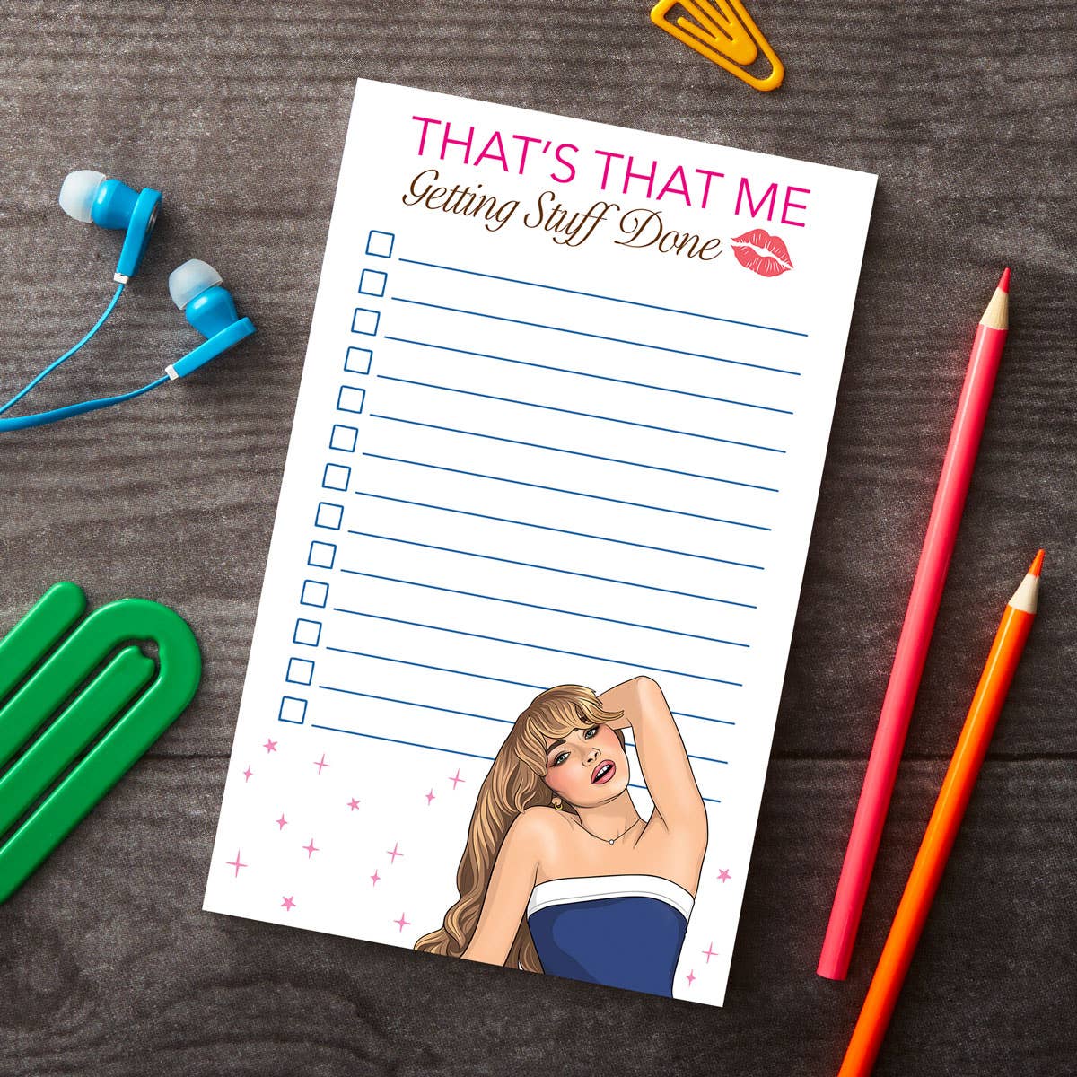 A notepad with a cartoon illustration of a female character in a blue dress, titled 'That's That Getting Stuff Done' with a pink lipstick mark on the cover, placed on a wooden surface alongside a pair of pencils and earphones.