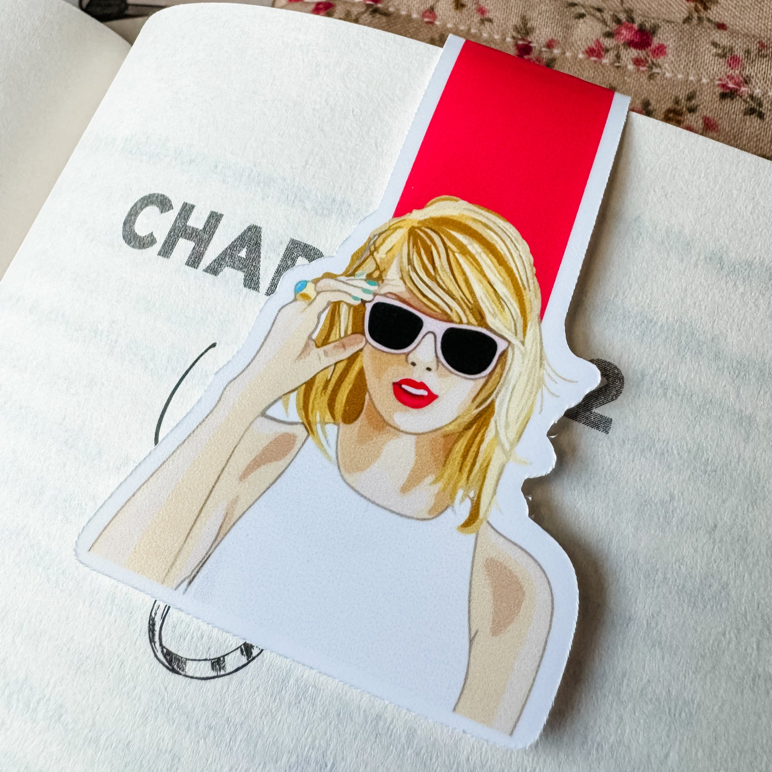 A colorful magnetic bookmark featuring an illustration of a person with blonde hair and sunglasses, predominantly white and red, placed on an open book.