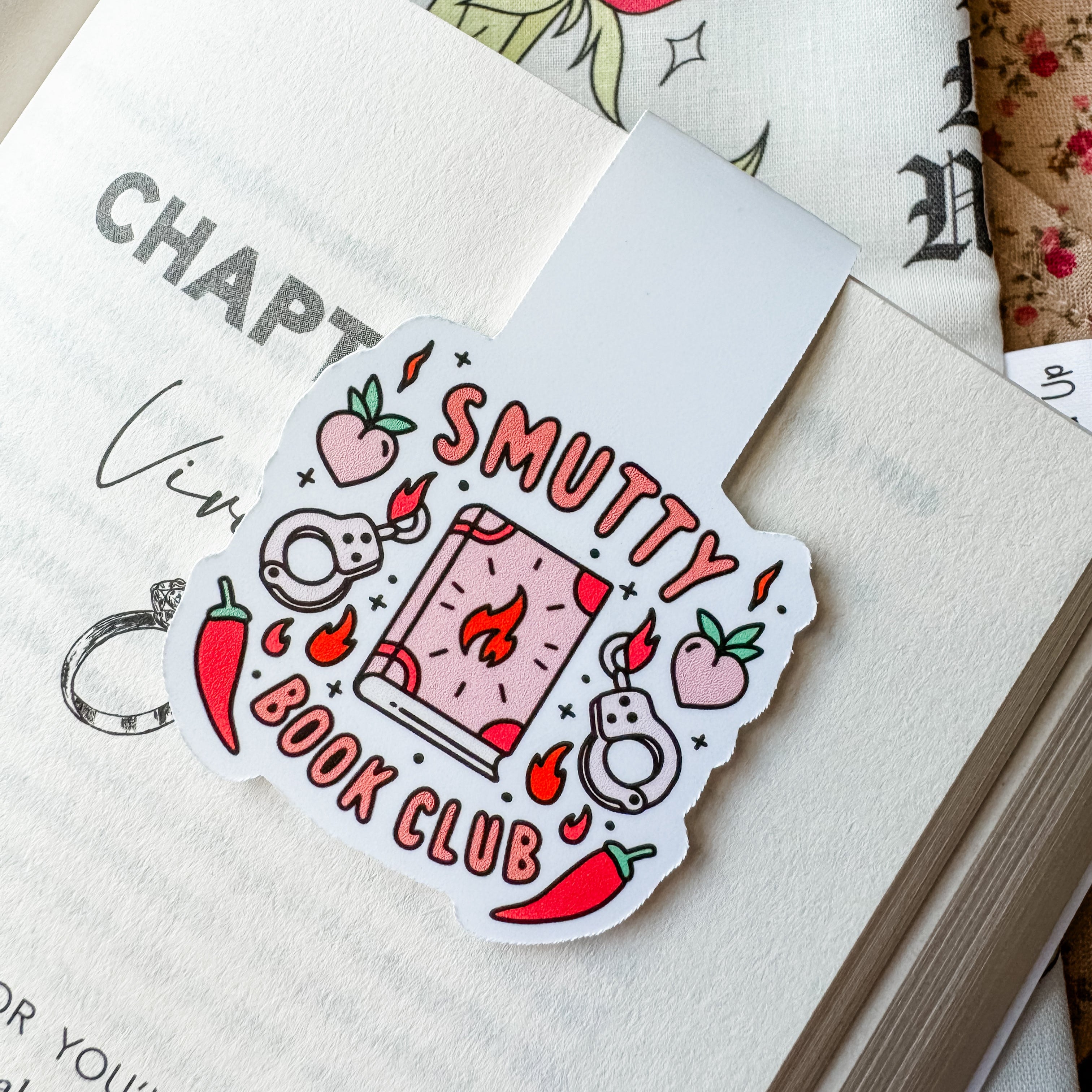 A magnetic bookmark with a colorful design featuring text and illustrations related to the 'Smutty Book Club', placed on an open book.