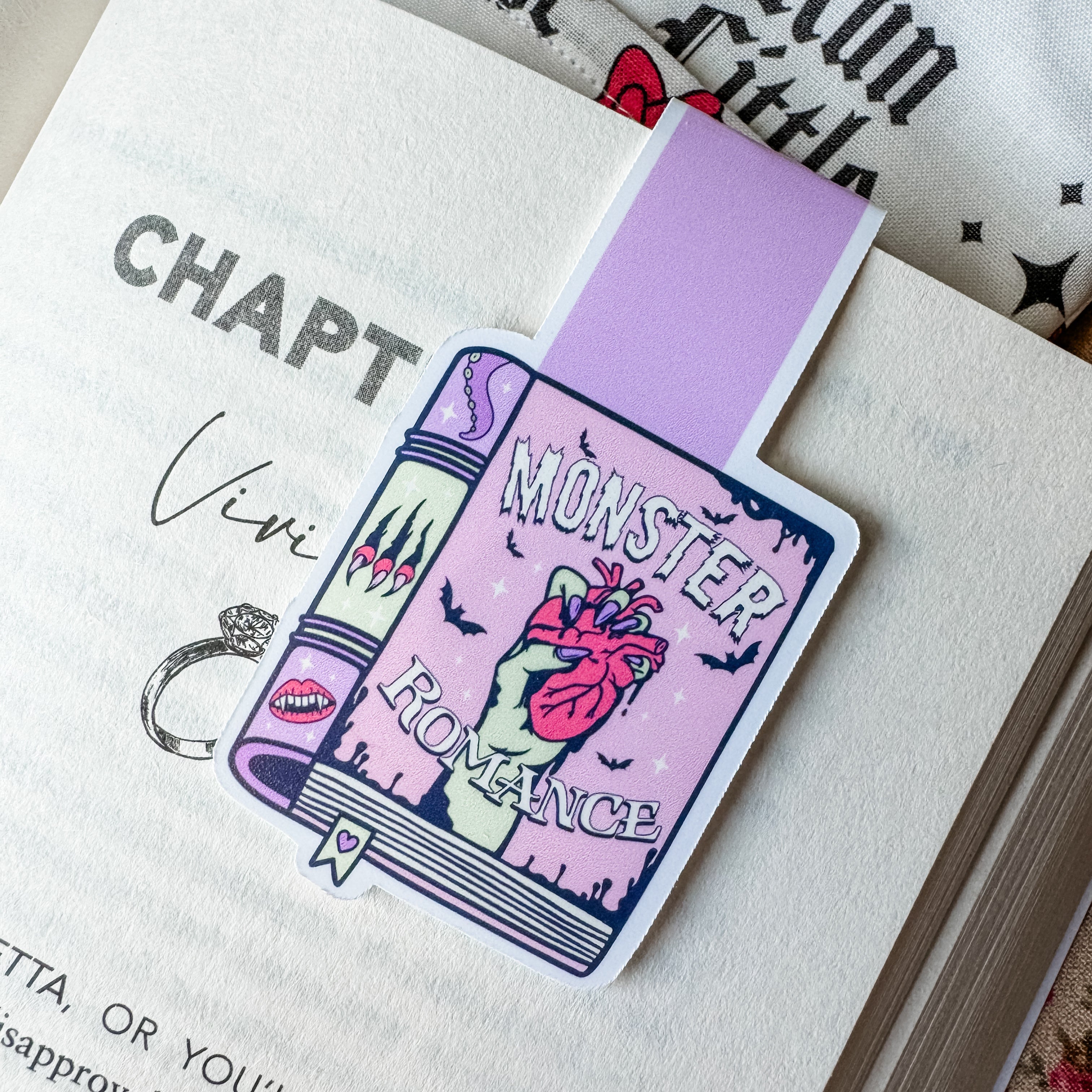 A magnetic bookmark with a colorful design featuring text and a romantic-themed monster illustration, placed on an open book.