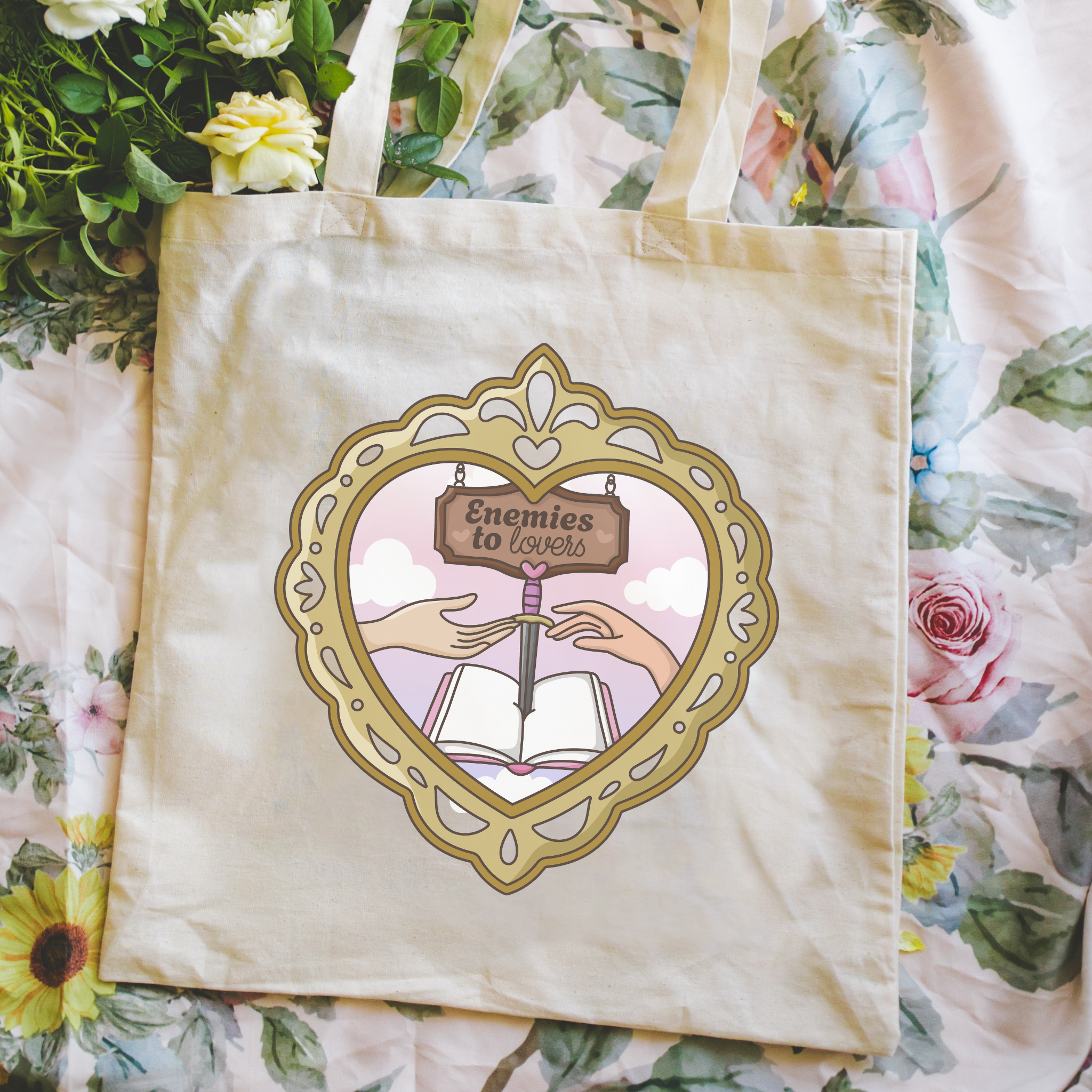 Tote bag with a decorative design featuring hands holding an open book and a heart-shaped frame, with 'Enemies to Lovers'  and text on a floral background