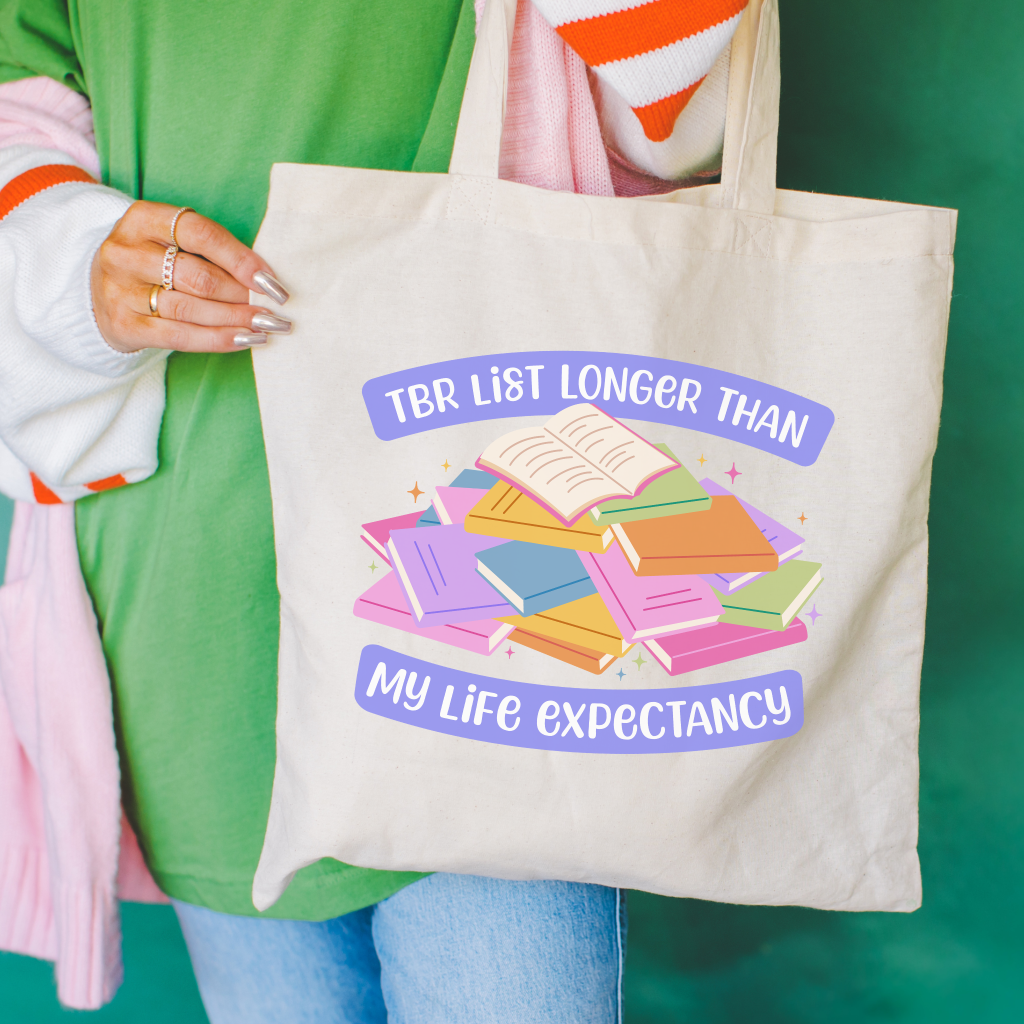 Bookish colorful tote bag with a pile of books and the text "TBR List Longer Than My Life Expectancy"held by a person against a green background