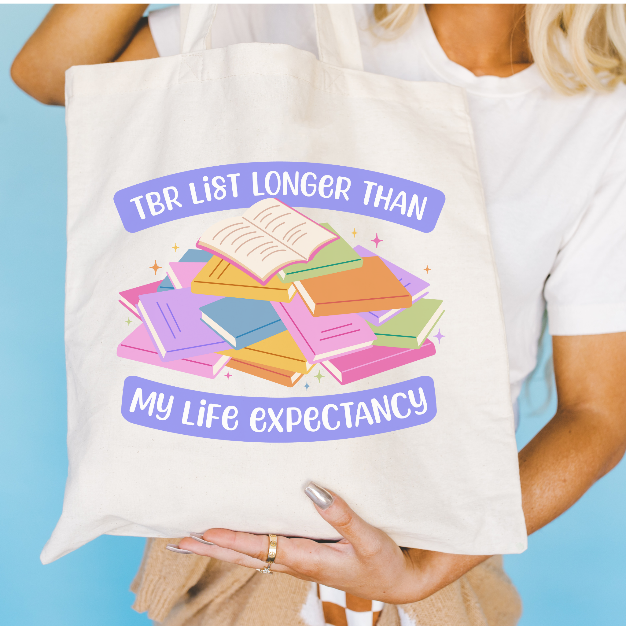 Bookish colorful tote bag with a pile of books and the text "TBR List Longer Than My Life Expectancy"held by a person against a blue background