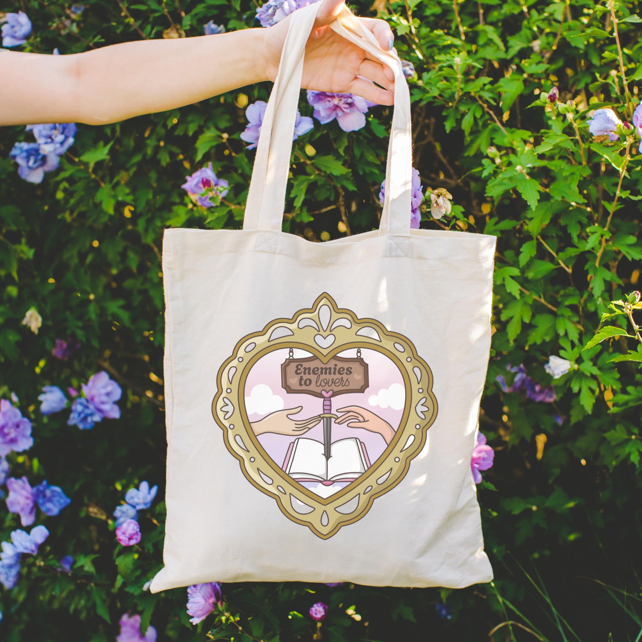 Tote bag with decorative design featuring hands, an open book, and "Enemies to Lovers" on a floral background