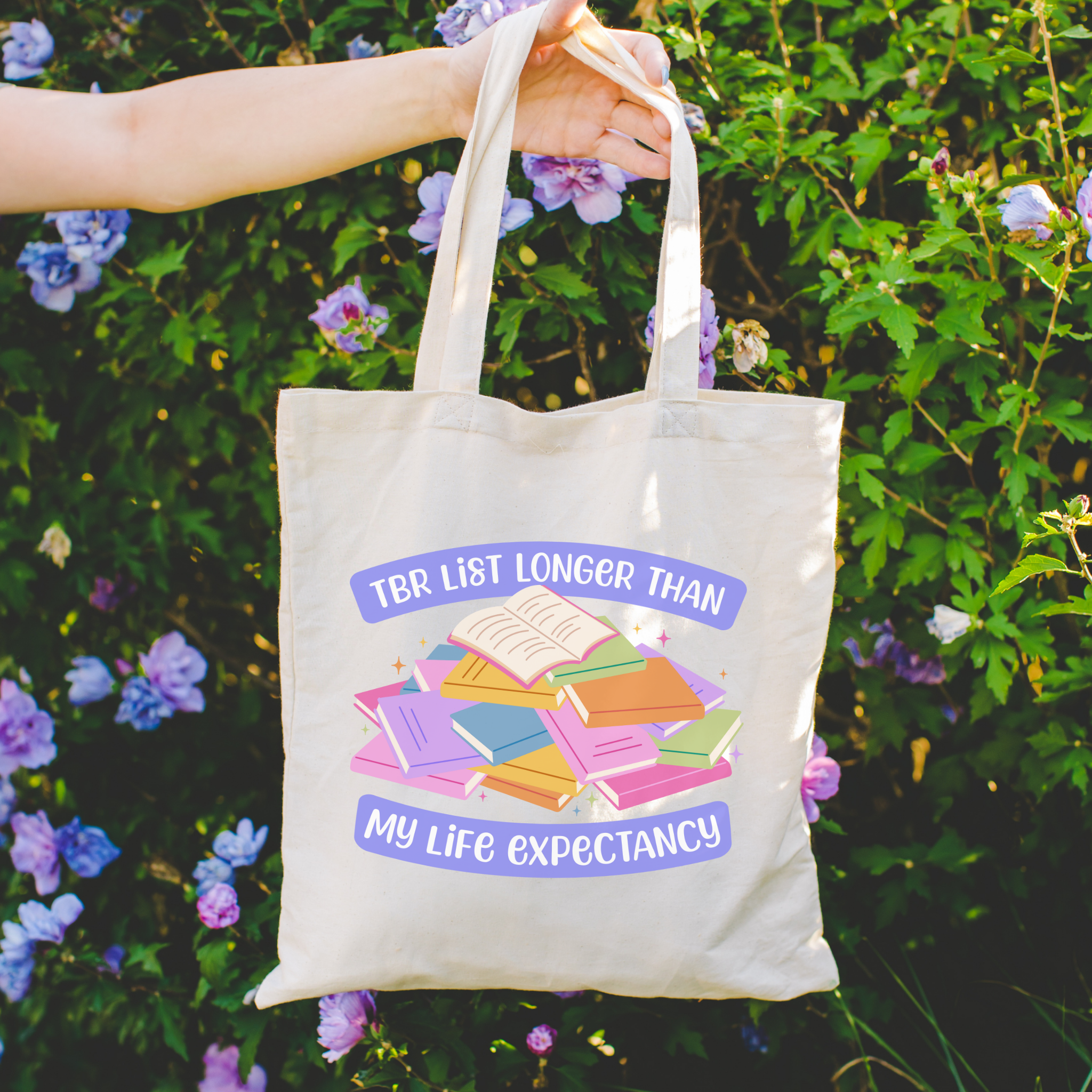 Bookish colorful tote bag with a pile of books and the text "TBR List Longer Than My Life Expectancy"held in front of a floral background