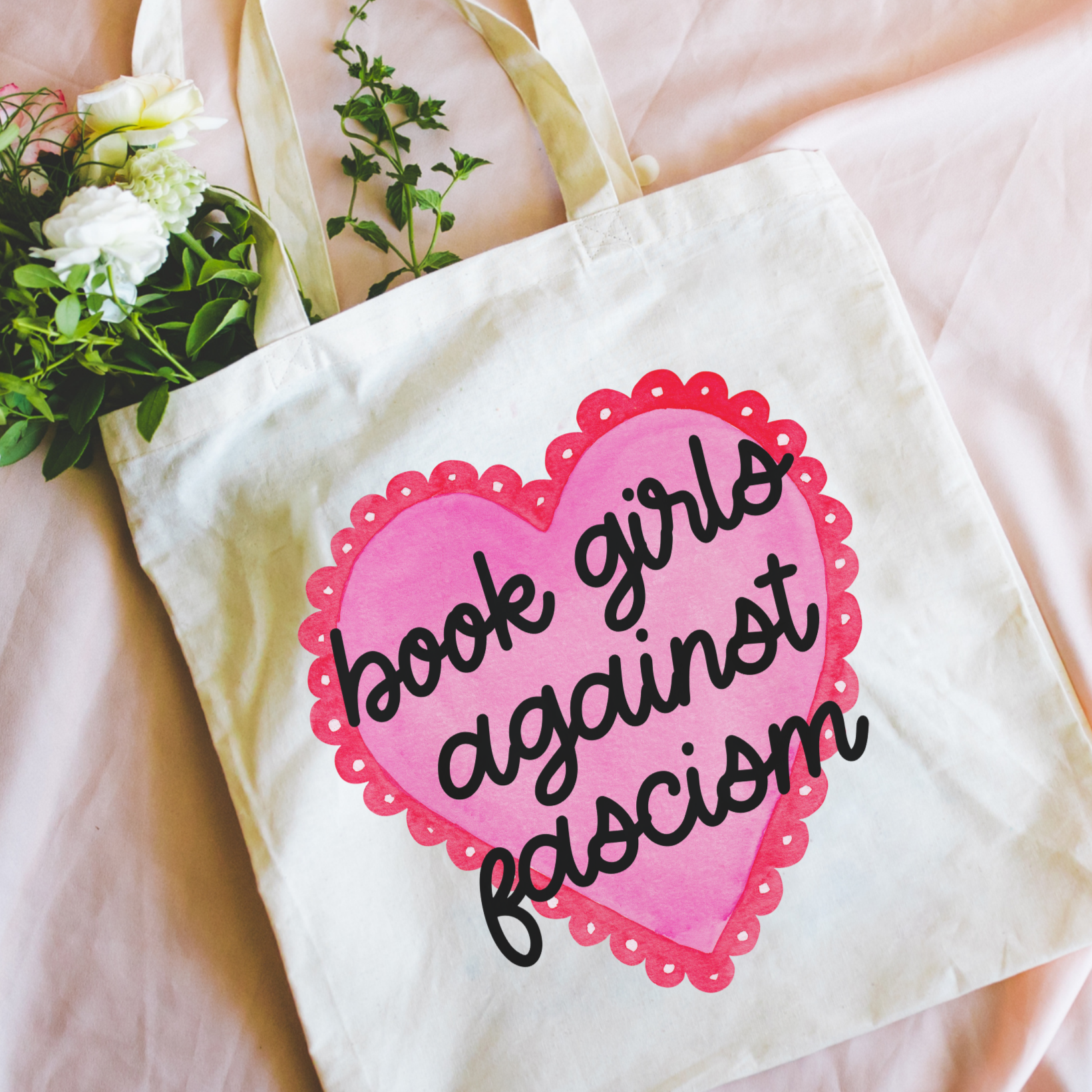 Bookish tote bag with a pink heart and text on a pink surface with flowers.