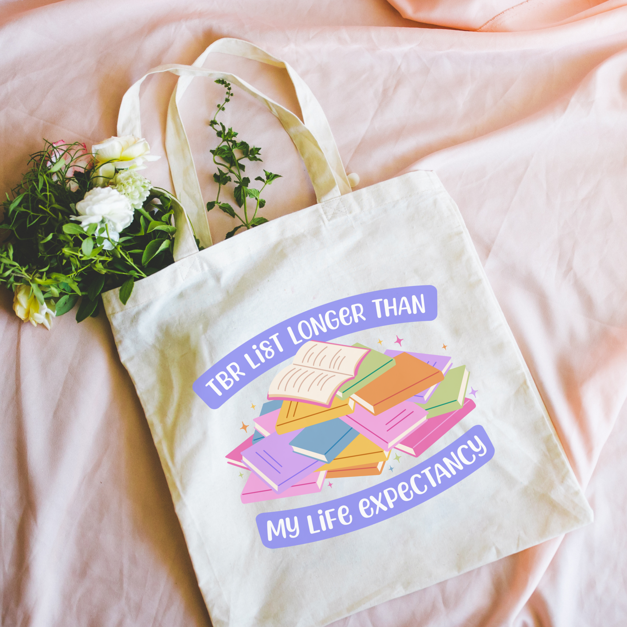 Bookish colorful tote bag with a pile of books and the text "TBR List Longer Than My Life Expectancy"  on a pink surface with flowers.