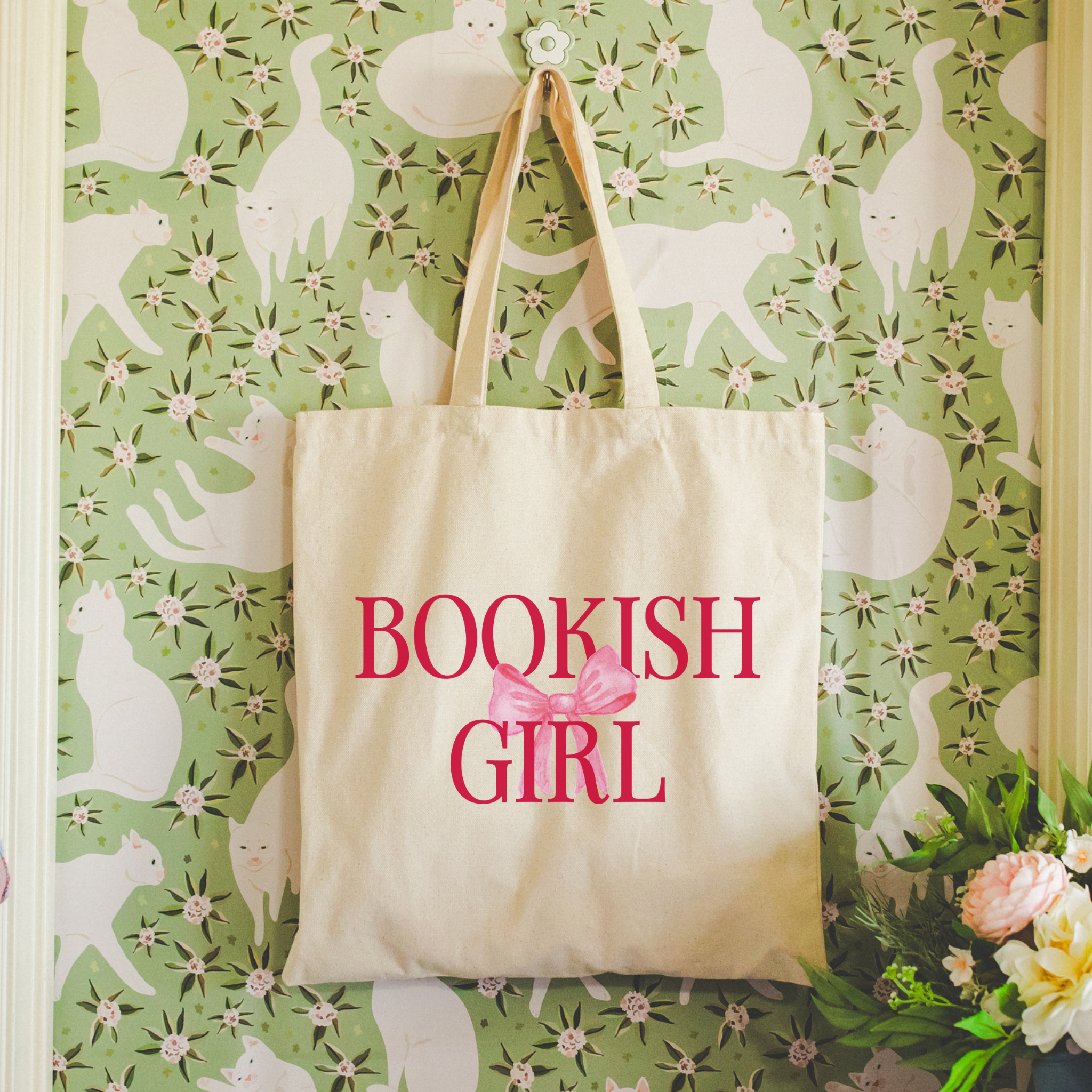 Beige tote bag with 'BOOKISH GIRL' text and pink bow against a floral cat-patterned wall.