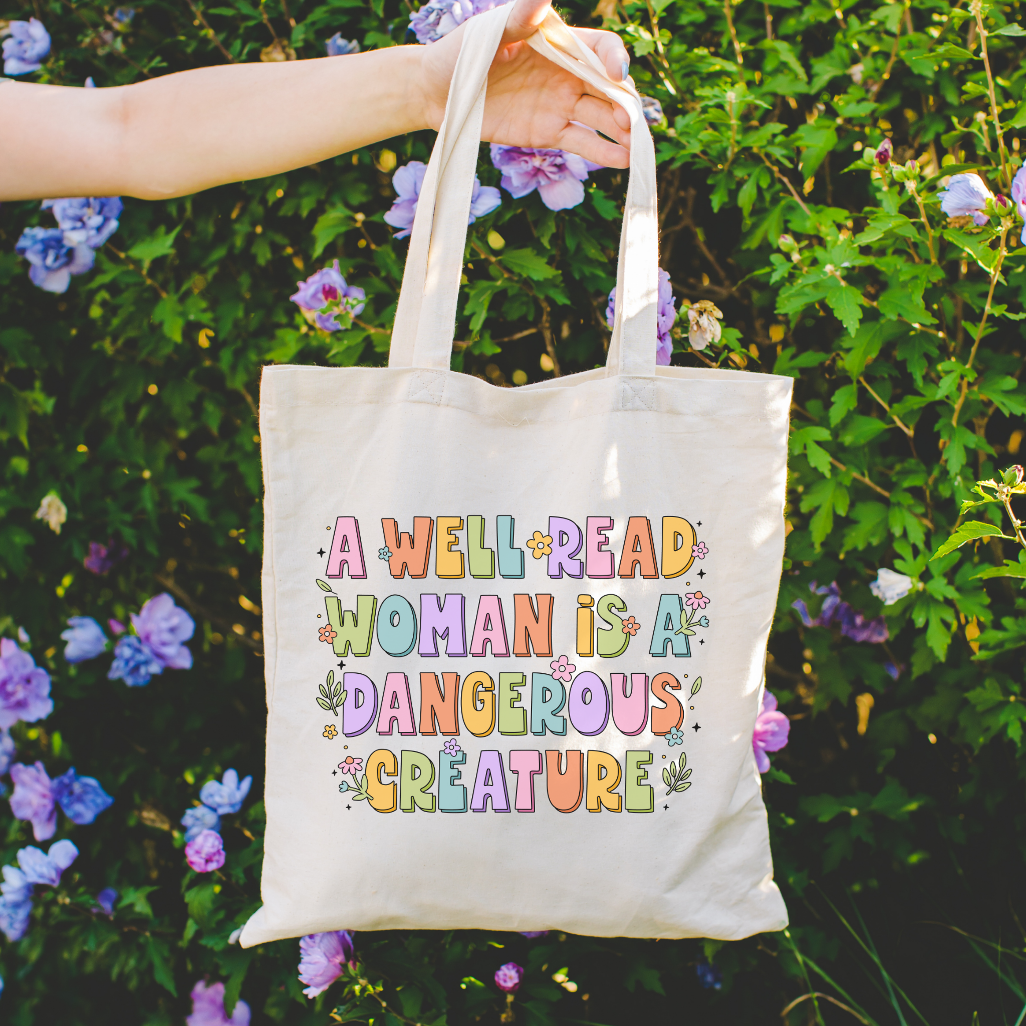Bookish Tote bag with colorful text held in front of a floral background and the text A Well Read Woman is a Dangerous Creature 