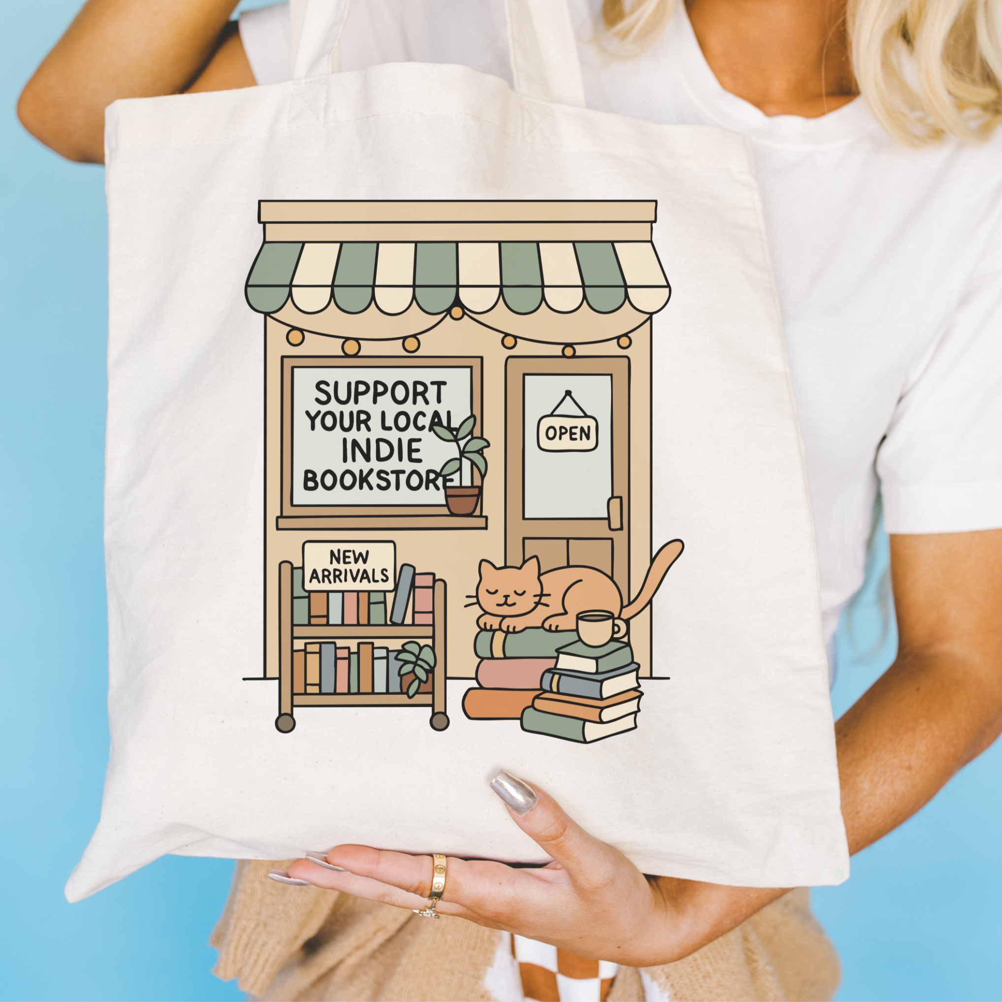 Tote bag with a design of a cat on books and a store front, held by a person against a blue background.