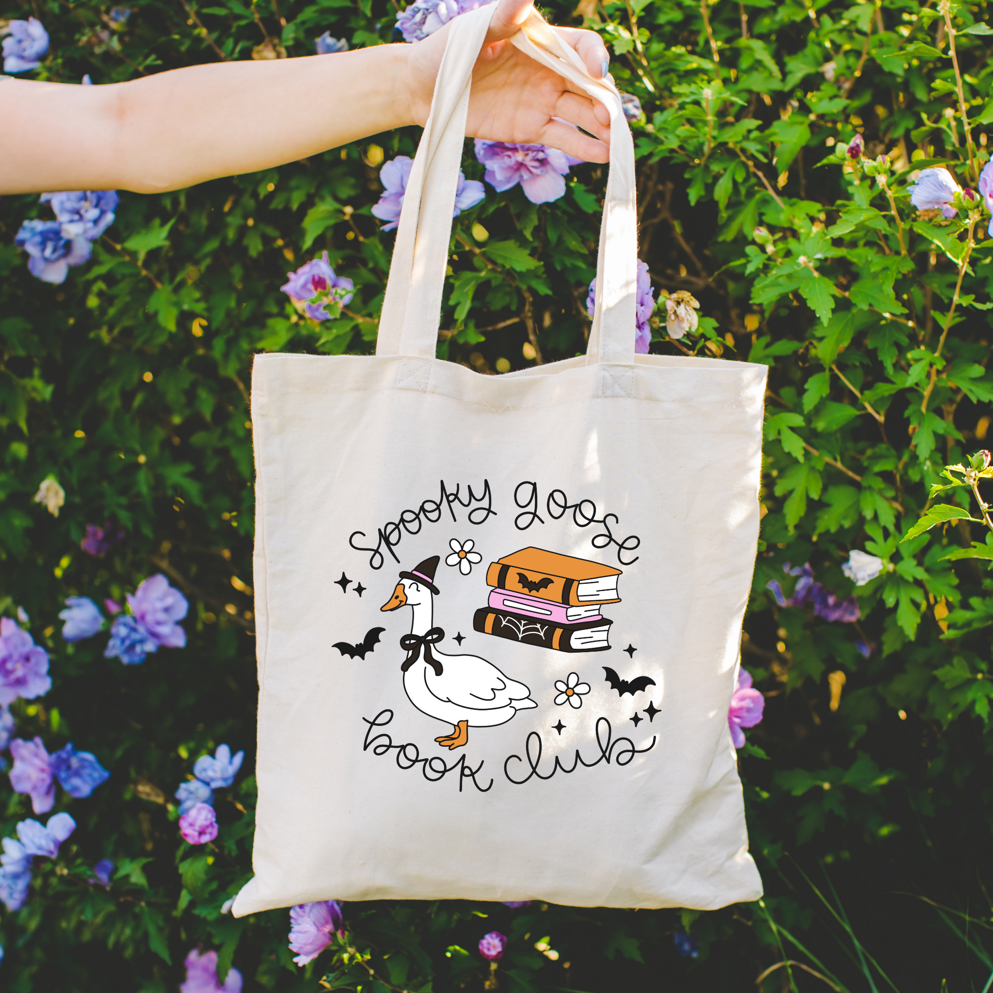 Tote bag with 'Spooky Goose Book Club' design held in front of a floral background