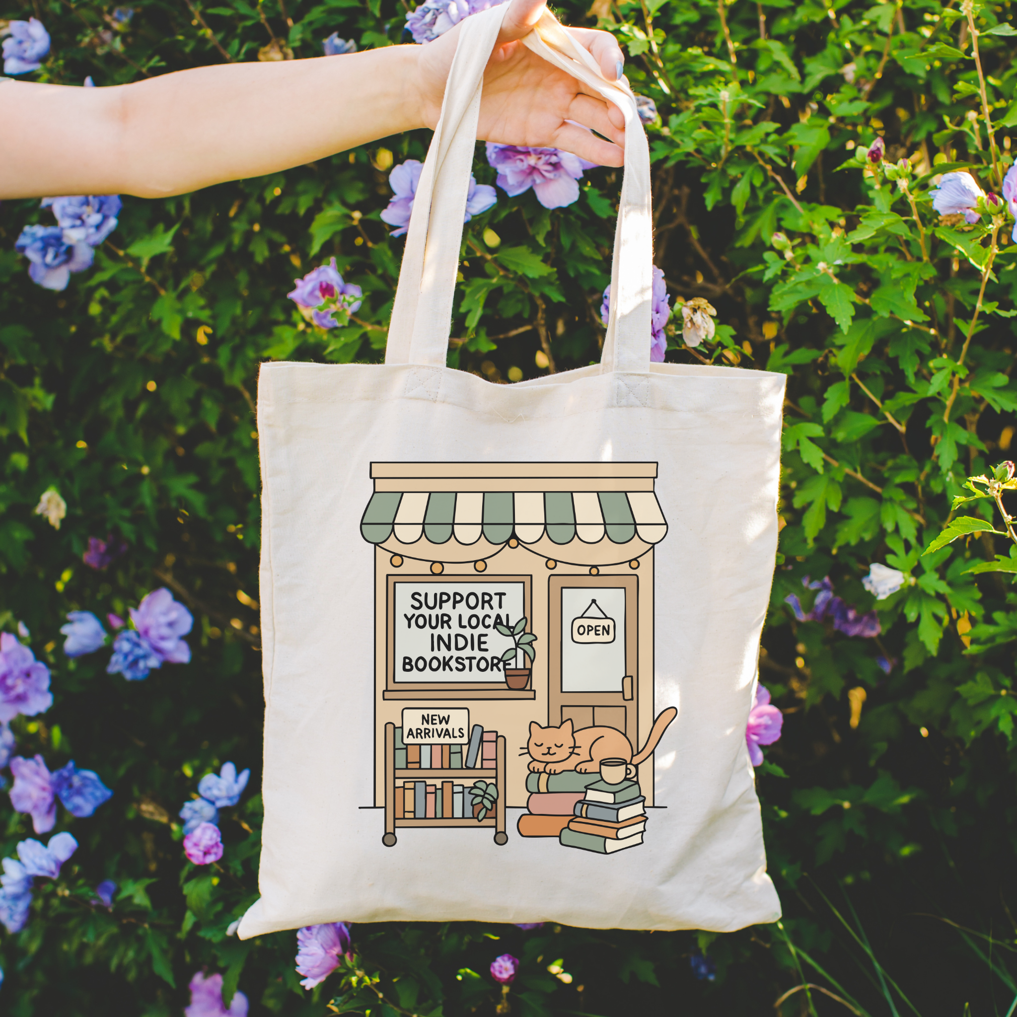 Tote bag with a design of a bookshelf, cat, and text 'Support Your Local Indie Bookstore' held against a floral background.