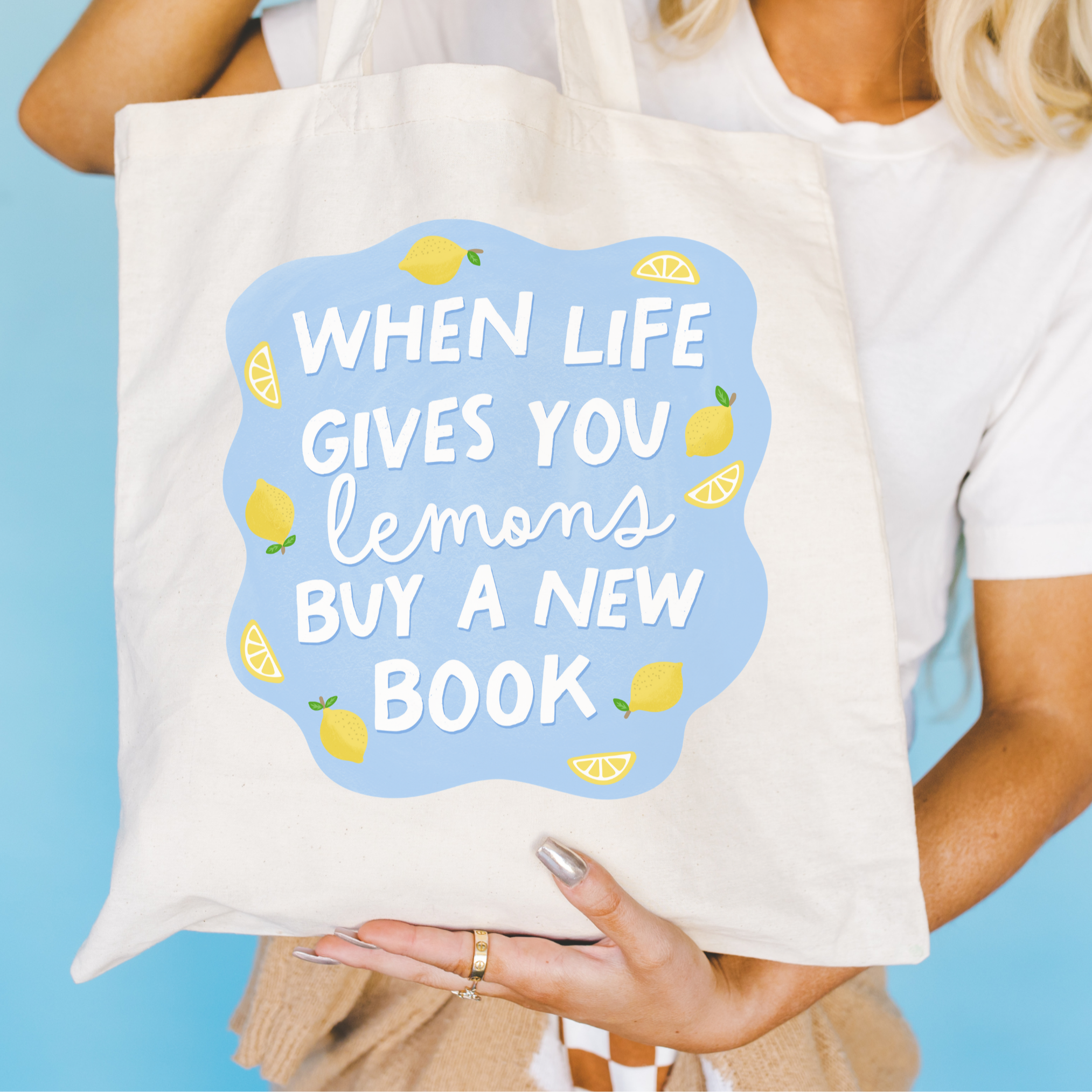 Tote bag with motivational quote 'When life gives you lemons buy a new book' held by a person against a blue background