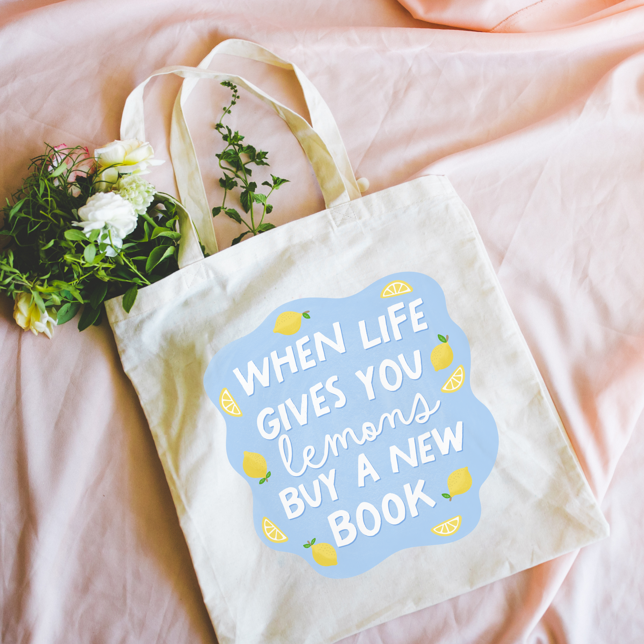 Tote bag with motivational quote 'When life gives you lemons buy a new book' held by a person against a pink background