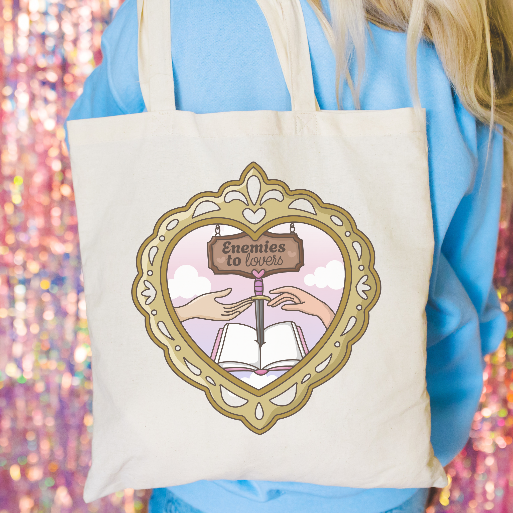 Tote bag with a decorative design featuring hands holding an open book and a heart-shaped frame, with 'Enemies to Lovers' text.