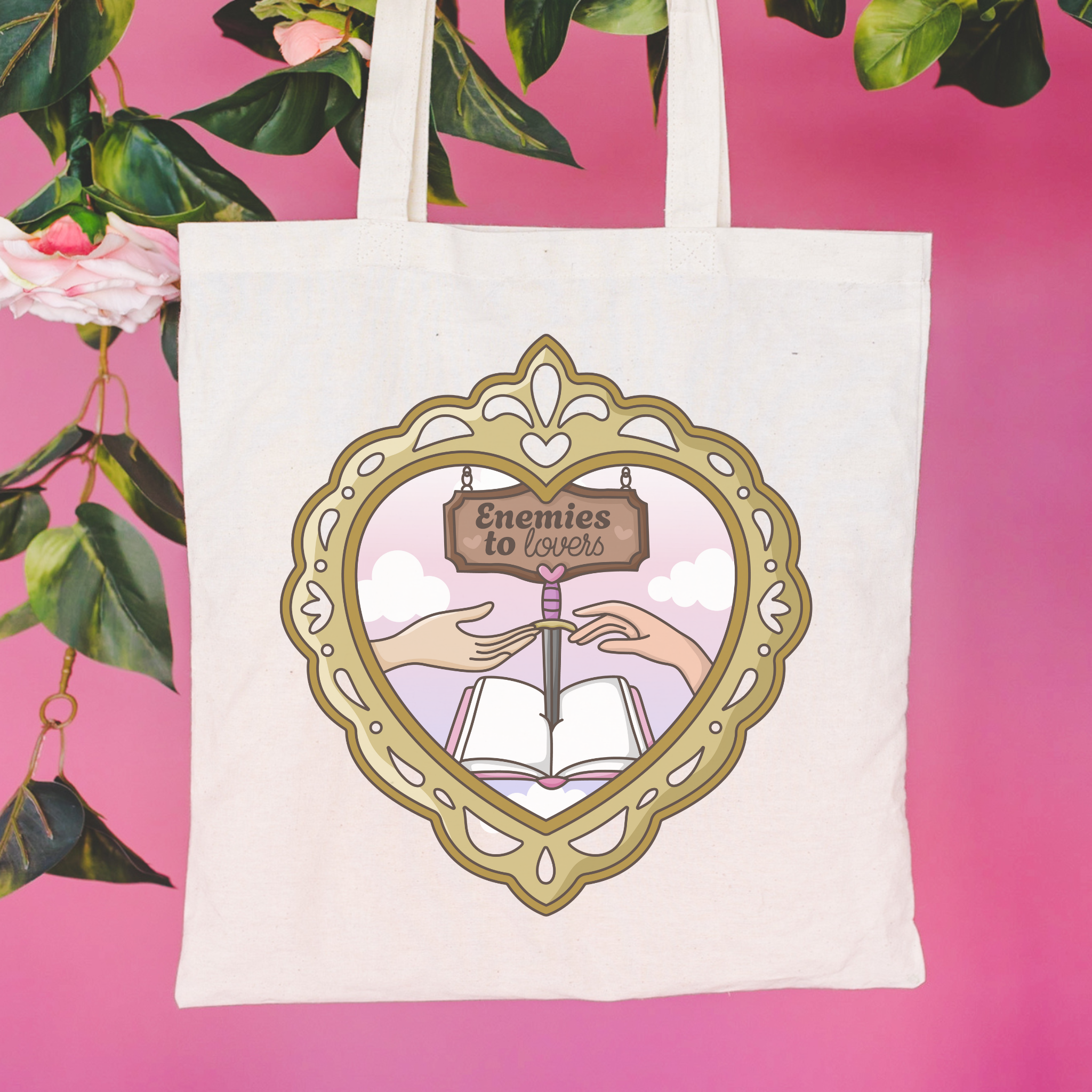 Tote bag with a decorative design featuring hands holding an open book and a heart-shaped frame, with 'Enemies to Lovers'  and text on a pink background