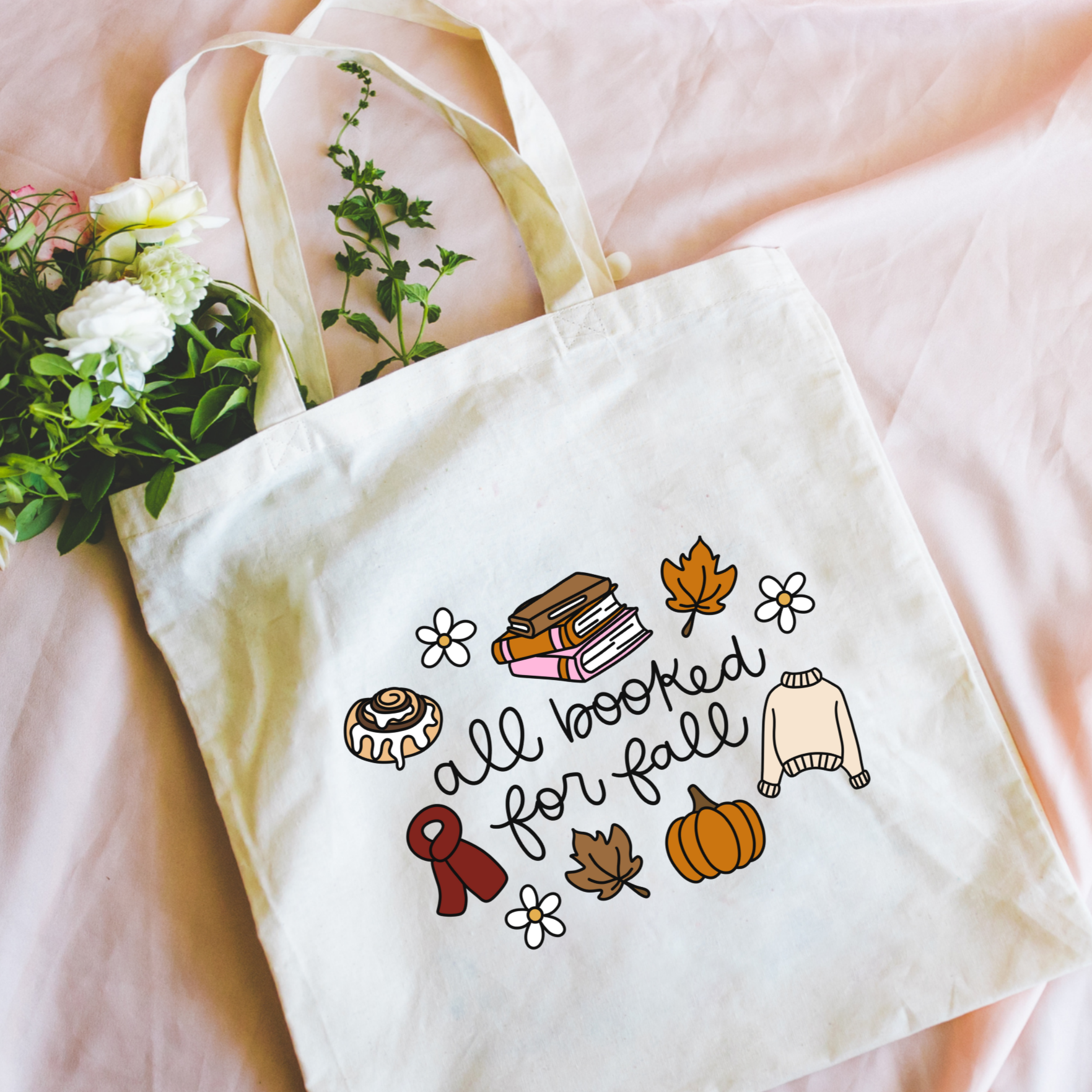 Bookish tote bag with autumn-themed illustrations and text on a pink surface with flowers.