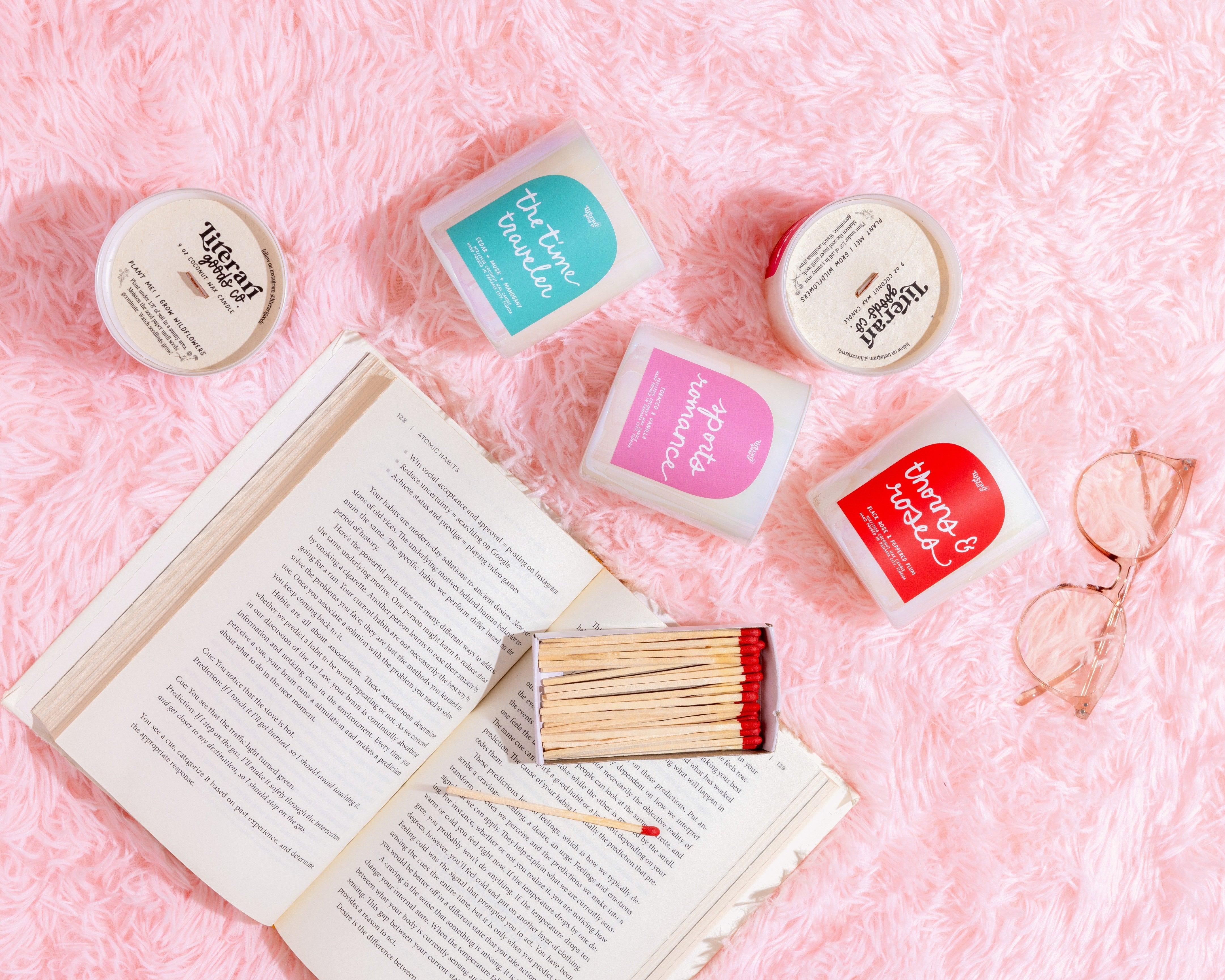 book inspired Candle tins with colorful labels on a pink textured surface next to an open book and matches.